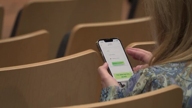 A woman uses a smartphone in an empty auditorium, reading Latvian text on Facebook, seated between light wooden chairs in a casual event setting.

