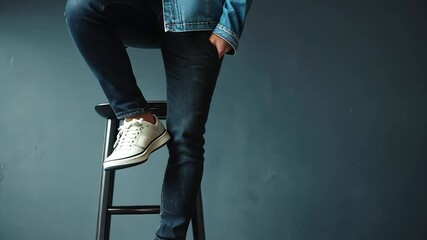 Man in denim jacket and dark blue jeans, posing with white sneakers on a black stool against a dark teal wall, showcasing casual fashion