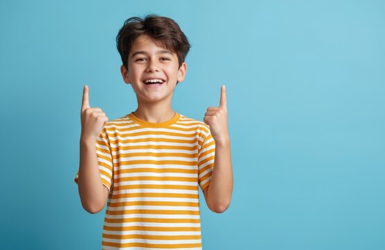 Young boy in striped t-shirt points up with index finger, shows direction, indicates choice on blue background with copy space. Happy child portrait, smiling face, positive emotions, joyful childhood.