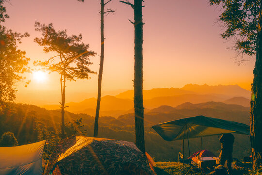 A man in winter clothing is standing on a campsite enjoying the morning light. The sun rises behind Doi Luang Chiang Dao mountain in Chiang Mai, Thailand. There are two tents and dense forest trees. - Powered by Adobe