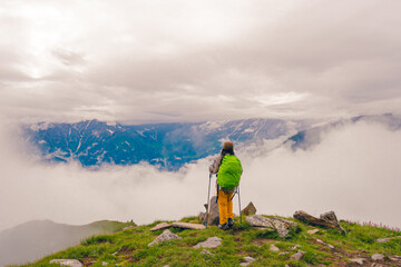Photo from the back of a female hiker standing at a high mountain pass surrounded by dramatic clouds and rocky alpine terrain. Idea for a solo trekking adventure in the Himalayas.