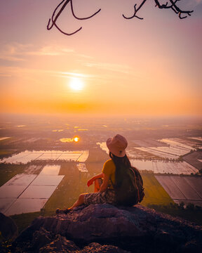Picture from the back of a man standing on the peak of high mountain cliff. There are a lot of rock with white color. The sun is setting on the mountain and there is a beautiful warm orange light.