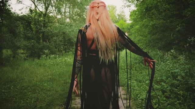 Halloween holiday woman witch walking in dark forest path green trees. long black dress gothic boho style macrame, elf fairy girl shaman back rear view. orange colored blonde hair braid hairstyle