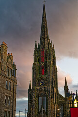 Fototapeta premium Gothic church in Edinburgh at sunset with clouds. Vertical view of beautiful bell tower of Tolbooth Kirk church in old town. Dark clouds with orange sun light.