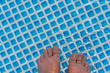 Two feet dip into the clear water of a pool. The sun's rays create light patterns on the skin and...