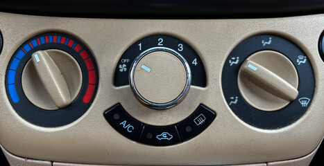 Close-up of a car climate control panel, showing temperature adjustment, fan speed, and air distribution settings. The dashboard features beige and black controls with red and blue color indicators
