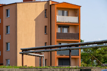Overground district heating pipes leading to a multi-family residential building with balconies. The block is painted in shades of beige and orange, with sky and trees in the background.