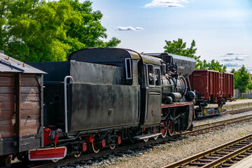 Obraz premium old, historic narrow-gauge steam locomotive with freight cars at the narrow-gauge railway station in Zbiersk, Greater Poland Voivodeship, surrounded by greenery on a sunny day.