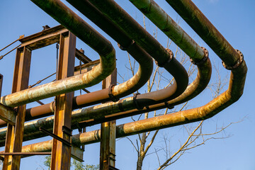 Rusty district heating pipes mounted on an overhead metal structure, with bare trees and blue sky...