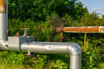 section of an insulated industrial pipeline with metal-clad valves running through a grassy, overgrown area. In the background, a rusty steel pipe contrasts with the green vegetation.