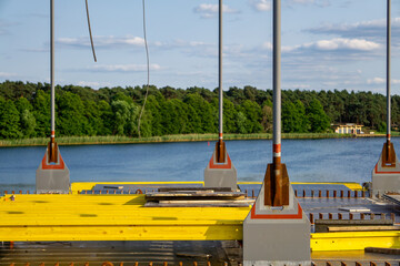 Section of a bridge under construction over water, with visible yellow formwork and metal support elements. A scenic background with a lake and green forest.