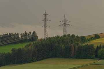 Electric towers stand tall over rolling green hills under a cloudy sky in a rural landscape during...