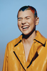 Laughing woman with colorful short hair wearing a bright yellow textured jacket against a soft blue...
