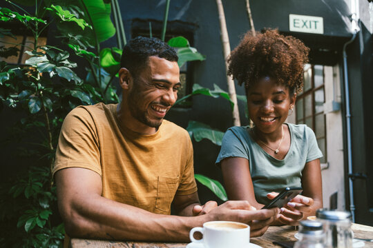 Happy couple using smartphone at table in cafe surrounded by plants