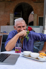attractive middle-aged man with white hair showing a red rose during an outdoor aperitif