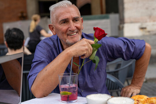 attractive middle-aged man with white hair showing a red rose during an outdoor aperitif