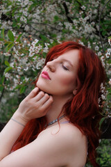 Fototapeta premium red-haired girl with curly hair walks in the park. Girl in a smart dress smiles. Woman poses against the background of blooming yellow dandelions and white flowers in a turquoise dress