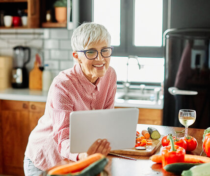 Portrait of happy senior mid aged mature woman prepering meal with fresh vegatebles and following internet instructions for a recipe on a tablet computer in kitchen