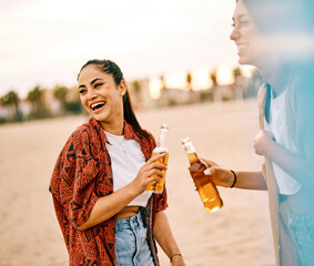 Portrait of two beautiful cheerful young women having fun drinking beer and toasting  on the beach....