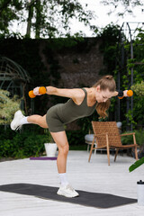 Athletic woman in green sportswear balancing on one leg while lifting dumbbells outdoors on a mat,...