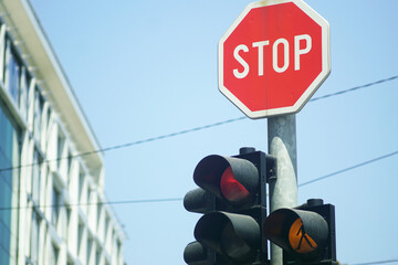 Red light signal and stop sign against backdrop of city buildings and blue sky. Urban mobility rules, transportation safety and traffic management. Modern street life and pedestrian alert concept