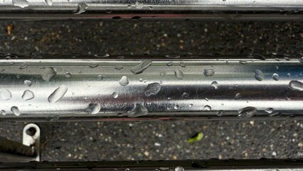 Water droplets on stainless steel metal surface with dark asphalt background, close-up of wet shiny texture, rain on polished steel pipes, minimal urban composition
