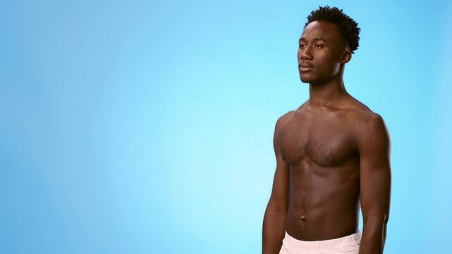 A young African American man stands tall and confident, showcasing his physique. The solid blue background enhances his presence, creating a striking visual impact, copy space