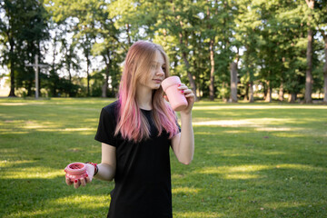 Fototapeta premium Teenage girl with pink hair drinks water from a reusable cup while exercising outdoors in a lush green park, promoting hydration and fitness