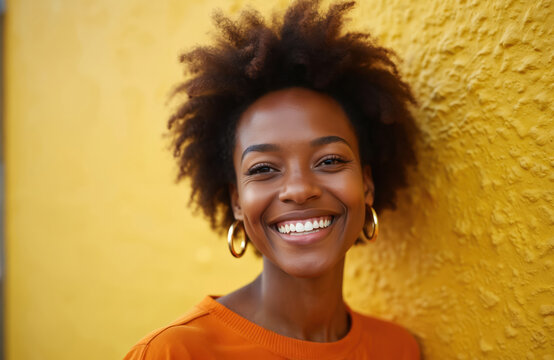 Portrait of radiant young black woman with natural afro hair smiles against textured yellow wall. Cheerful adult female with hoop earrings looks forward with confidence, positivity. - Powered by Adobe