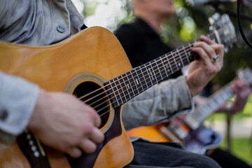 Fototapeta premium close up of acoustic guitar player performing live music outdoors with band, fingers strumming strings, musicians playing instruments in park, street performance, live acoustic session background