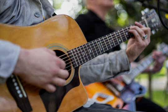 close up of acoustic guitar player performing live music outdoors with band, fingers strumming strings, musicians playing instruments in park, street performance, live acoustic session background