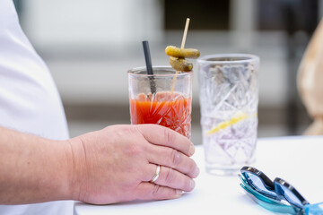 man holding bloody mary cocktail in crystal glass with olives and straw on white table at outdoor...