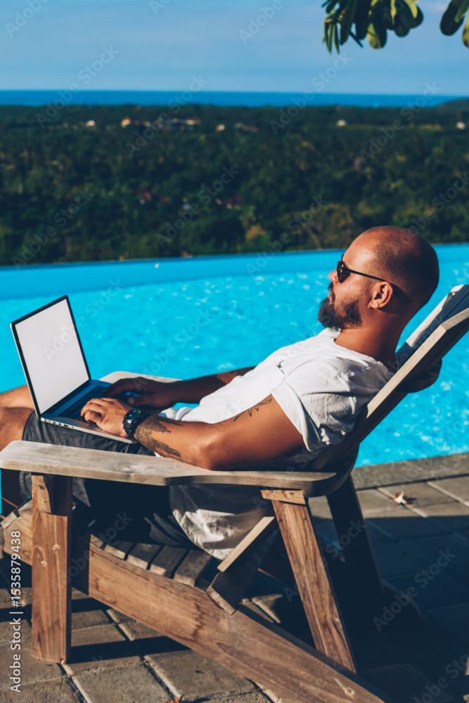 Poster bearded businessman lying near blue pool while typing text on keyboard of netbook with blank copy sp