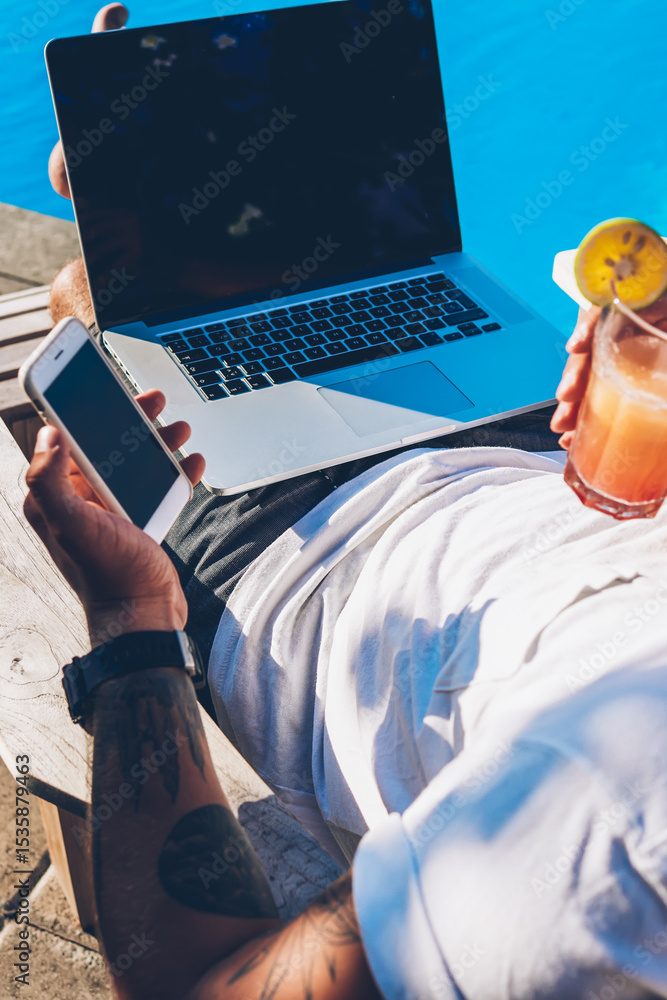 Poster young businessman with cocktail in hand reading notification on smartphoe while resting on sunbed ne