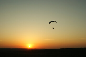 A paraglider floats gracefully above a serene landscape at sunset, with warm hues painting the sky as the sun descends behind the horizon. The atmosphere is calm and inviting.