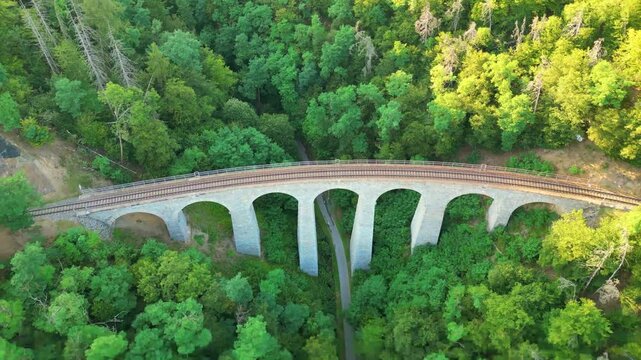 This stunning aerial view showcases the Zampach railway bridge, the highest stone bridge in Czechia, surrounded by lush greenery and smooth railway tracks through nature.