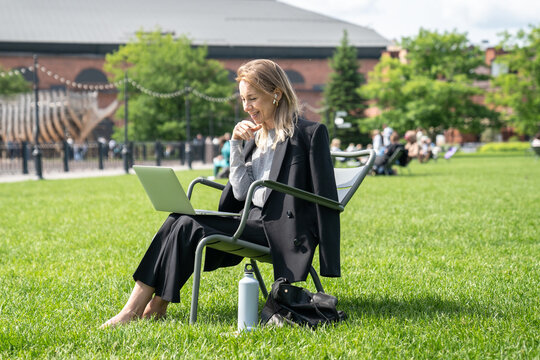 Laughing woman in earmuffs watching movie or video call on laptop in park on summer break. Female online entertainment outdoors, remote connectivity, leisure time, flexible work, urban park relax