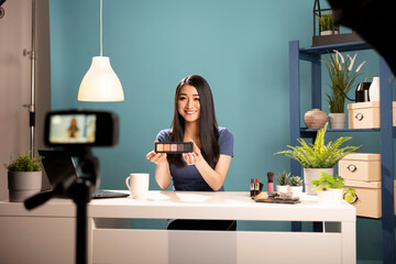 Female vlogger explains cosmetic products in a livestream video. Asian blogger holding eyeshadow palette, seated at a desk in studio with camera on tripod, creating engaging content for social media.