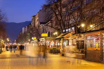 Night scene on Vitosha Boulevard in Sofia with lively cafes adorned with string lights, blurred pedestrians walking past glowing streetlights, and stunning view of Vitosha Mountain in background