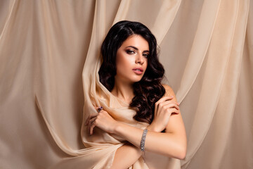 Elegant brunette woman posing against a beige draped background, showcasing beauty, elegance
