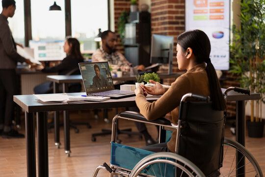 Female manager with a disability sitting in modern workspace, conducting a virtual meeting with a remote coworker. Asian woman in wheelchair with laptop, having a discussion with client on video call.