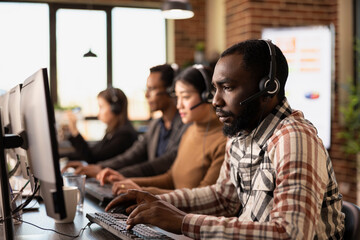 Portrait of african american customer support agent at his computer with coworkers nearby. Focused...