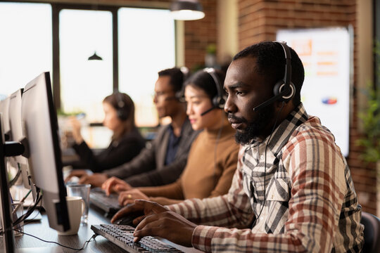 Portrait of african american customer support agent at his computer with coworkers nearby. Focused call center operator typing on his keyboard while listening to a client through his wired headphones. - Powered by Adobe