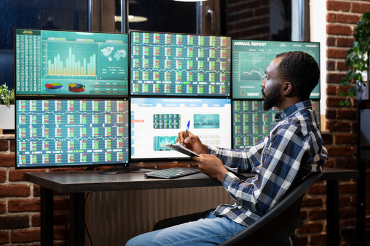 African american forex trader studies live market data on multiple monitors, taking notes on his clipboard in modern workspace. He remains focused during a trading session at tech savvy workstation.
