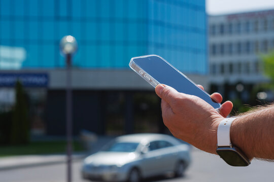 Close-up of a man hand holding a modern smartphone against a mirrored blue glass building in a city. Concept of technological lifestyle in urban environment.