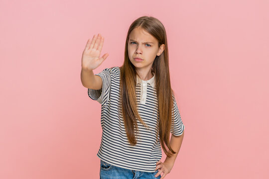Young child girl firmly raises one hand forward in stop gesture staring seriously into camera as if demanding to end prevent something with confidence clarity. School kid isolated on pink background