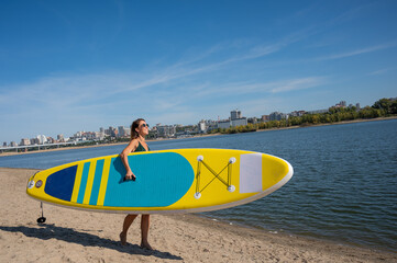 Caucasian woman walks along the beach and carries a sup board on the river in the city. Summer...