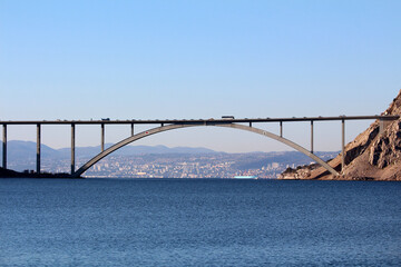A tall concrete arch bridge spans a wide blue bay, connecting rocky hillsides with a cityscape and distant mountains in the background, under a clear sky with vehicles crossing the bridge