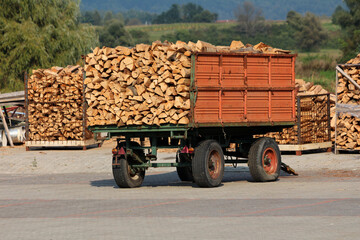 A rusty farm trailer with red metal sides is fully loaded with split firewood, parked on a paved...
