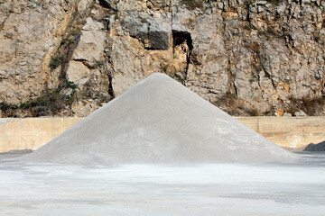 Large conical pile of fine gravel sits on a flat surface in a quarry, backed by a steep rocky wall,...